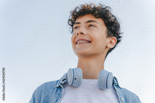 Portrait of smiling curly haired teenager with braces wearing headphones looking away standing on the street. Inspiration, technology concept   photo