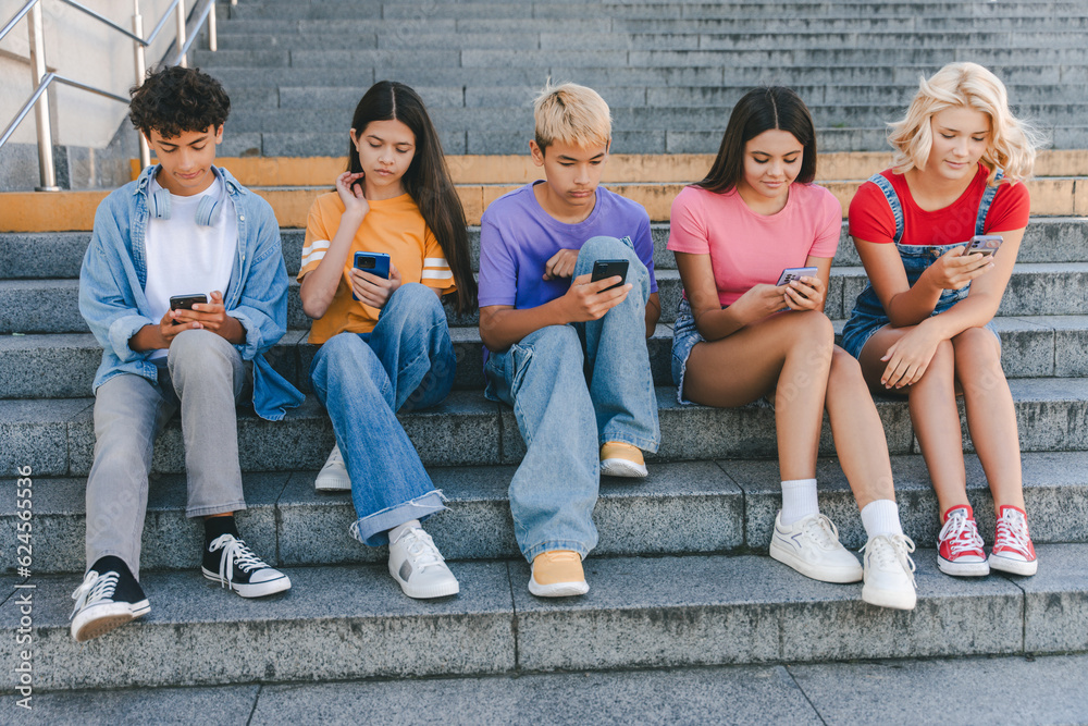 Obraz premium Group of serious friends, multiracial teenagers holding mobile phones watching video, chatting, communication online sitting on stairs. Technology addiction, social media concept