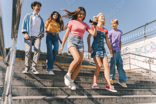 Fototapeta Group of smiling friends, multiracial teenagers wearing colorful casual clothes running on the street, having fun