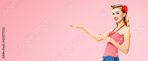 Image of smiling woman pointing at something. Excited girl in pin up, showing, holding some product or copy space area. Retro fashion and vintage. Rose pink color background.