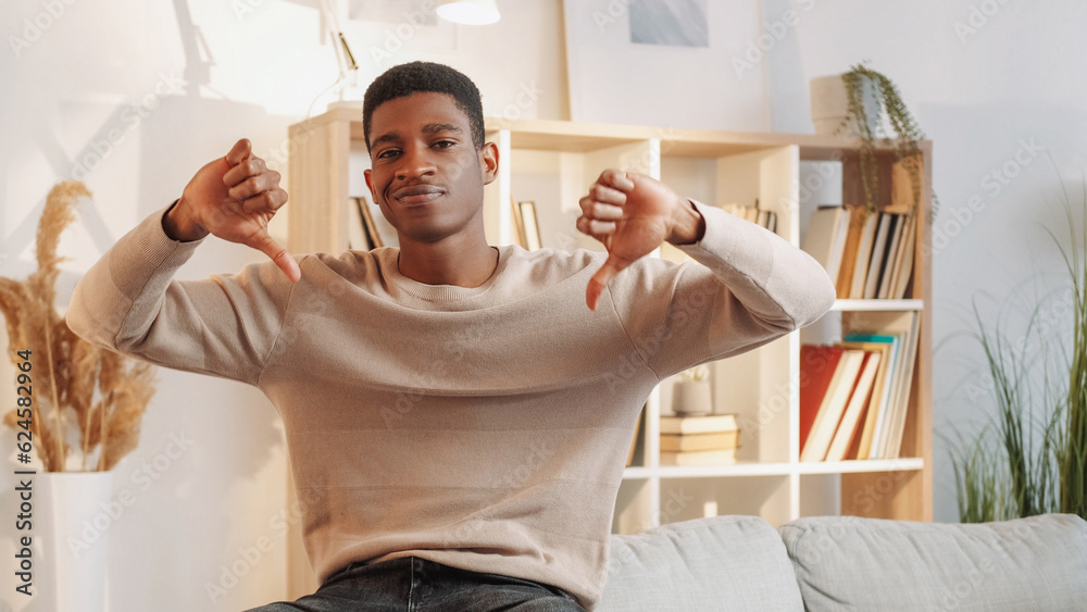 Obraz premium Bad recommendation. Thumbs down. Disappointment sign. Frustrated young man showing dislike gesture dissatisfied with house rent on couch at home living room.