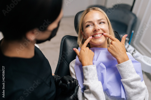 Close-up top view of happy young woman patient pointing with fingers at healthy smile, demonstrating whitening teeth procedure result, satisfied with dental clinic service. Concept of dental treatment © dikushin