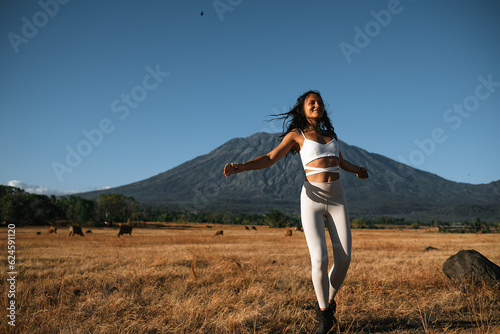
Portrait of a young, full of health girl, of European appearance, in a white yoga suit, posing for the camera, dancing, flying hair, against the backdrop of a mountain in a field with dry grass.