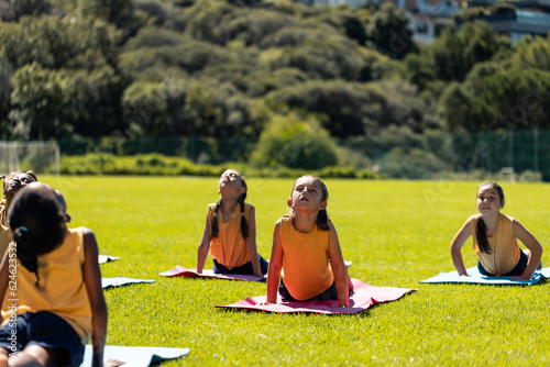 Diverse schoolgirls practicing yoga meditation on sunny elementary school sports field