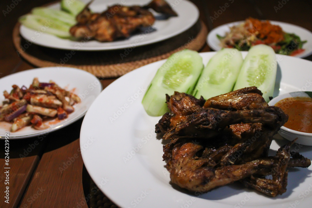 Ayam taliwang or taliwang chicken, a traditional roasted chicken dish from Lombok usually served with beberuk and water spinach plecing. Served with white plate on a wood background Ayam taliwang or taliwang chicken, a traditional roasted chicken dish from Lombok usually served with beberuk and water spinach plecing. Served with white plate on a wood background