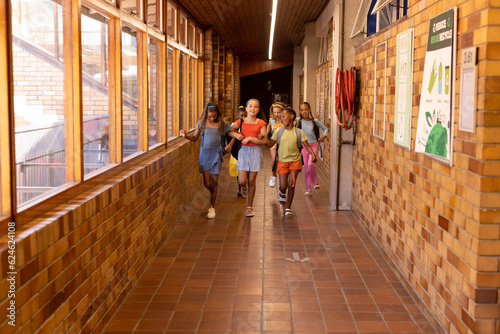 Diverse happy schoolgirls with bags running and embracing in elementary school corridor