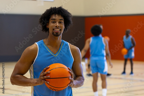 Portrait of happy biracial male basketball player wearing blue sports clothes over teammates at gym