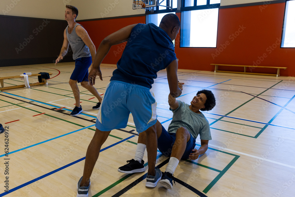 Diverse male basketball players helping to get up at gym