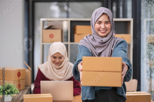 A happy young Asian Muslim female online seller working with her employee in the office. SME business, online shop owner