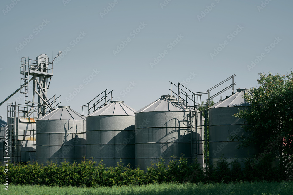 Steel grain silos stand next to a field. Agro-Industrial landscape. Steel Silos Embrace Vast Fields, Serving as Epitomes of Agricultural Storage and Processing Steel grain silos stand next to a field. Agro-Industrial landscape. Steel Silos Embrace Vast Fields, Serving as Epitomes of Agricultural Storage and Processing