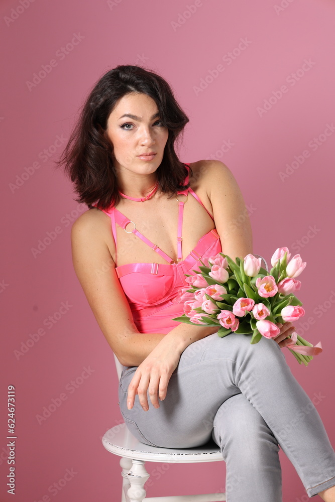 Pretty young brunette woman sit on chair with bucket of pink tulips. Girl in pink top and jeans with flowers smile. Woman barefoot, pink background. Portrait of beautiful woman