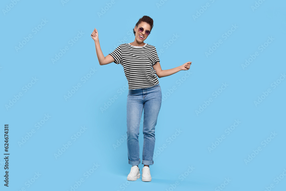 Happy young woman in stylish sunglasses dancing on light blue background Happy young woman in stylish sunglasses dancing on light blue background