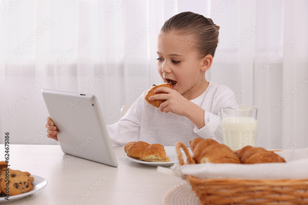 Little girl using tablet while having breakfast at table indoors. Internet addiction Little girl using tablet while having breakfast at table indoors. Internet addiction