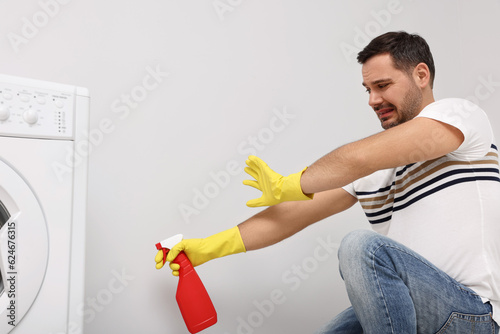 Emotional man in rubber gloves cleaning wall with sprayer indoors