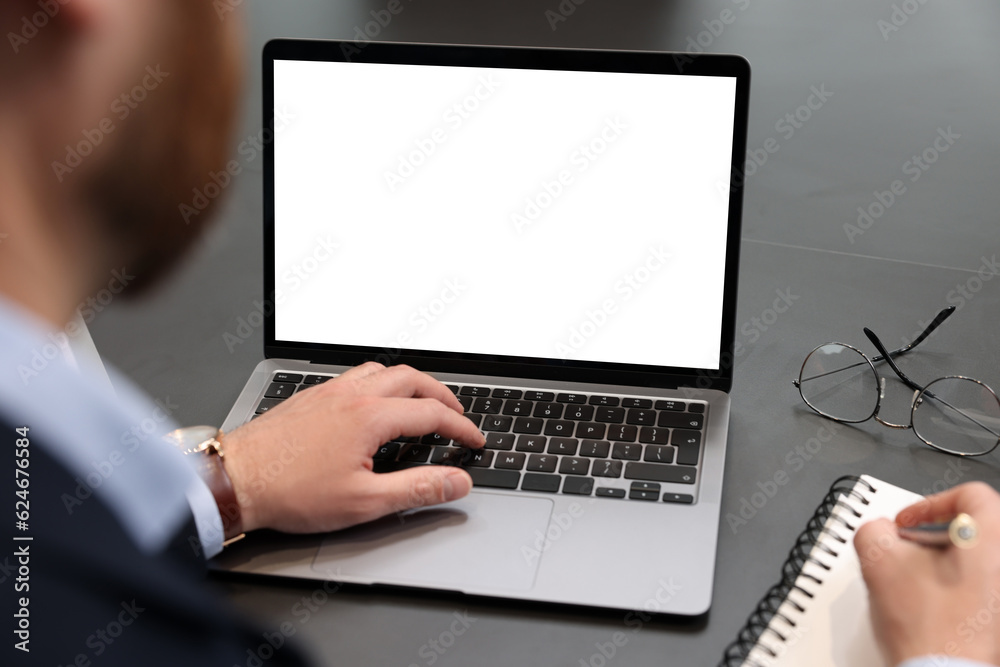 Man writing notes while working on laptop at black desk in office, closeup Man writing notes while working on laptop at black desk in office, closeup