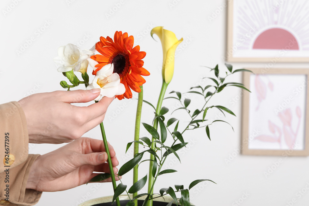 Stylish ikebana as house decor. Woman creating floral composition with fresh flowers near white wall with pictures, closeup