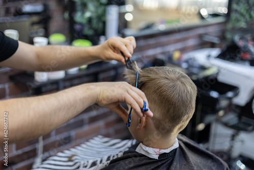 The hairdresser cuts the hair on the child's head with scissors. The boy sits in a chair in a beauty salon and watches the haircut process. The concept of a child in a barbershop.