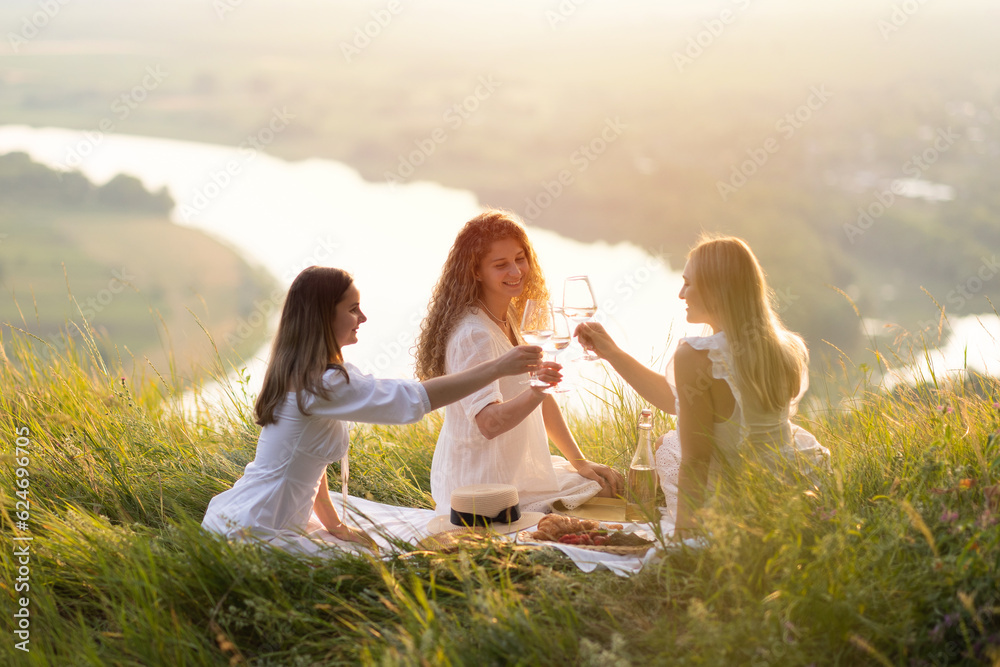 Three beautiful young caucasian ladies with wine in their hands enjoy spending time on picnic at summer sunset. Good weekend concept with friends