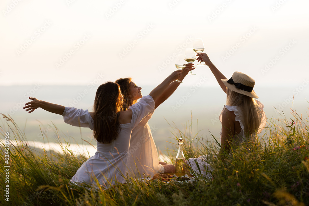 Three young beautiful smiling female in summer white dresses. Carefree women making picnic in the park at sunset. Positive models sitting on plaid on grass, drinking wine, eating fruits and cheese.