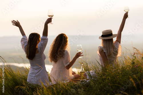 Obraz na płótnie Back view of girlfriends in stylish summer clothes raising arms with wine at sunset during picnic in summer park-
w salonie
