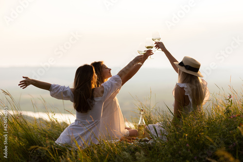 Obraz na płótnie Three young beautiful smiling female in summer white dresses. Carefree women making picnic in the park at sunset. Positive models sitting on plaid on grass, drinking wine, eating fruits and cheese.
w salonie