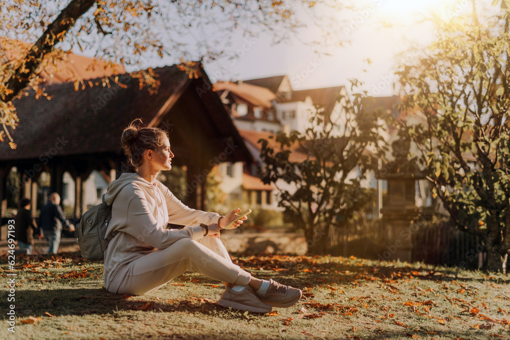 Obraz premium Young Fashionable Teenage Girl With Smartphone In Europian Park In Autumn Sitting At Smiling. Trendy Young Woman In Fall In Park Texting. Retouched, Vibrant Colors. Beautiful Blonde Teenage Girl