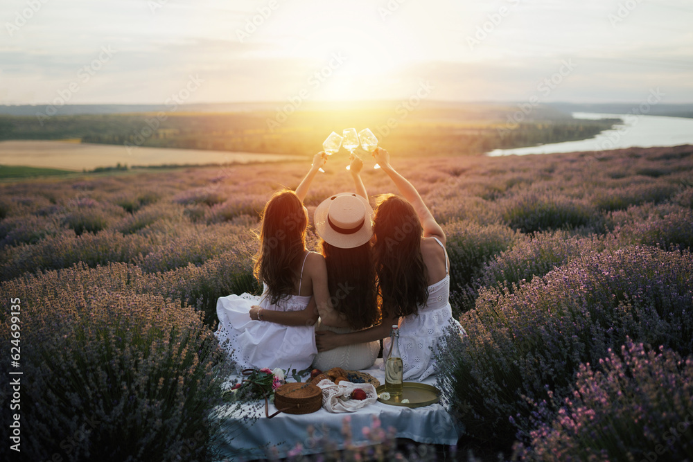 Girlfriends having picnic in the lavender field at sunset. Group of young women sitting on lavender field on summer day. Girlfriends drinking wine on outdoor party.