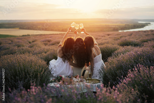Young female friends having fun, raising glasses with wine and enjoy beautiful sunset at summer picnic in lavender field.	