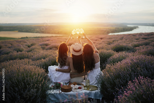 Obraz na płótnie Girlfriends having picnic in the lavender field at sunset. Group of young women sitting on lavender field on summer day. Girlfriends drinking wine on outdoor party. w salonie