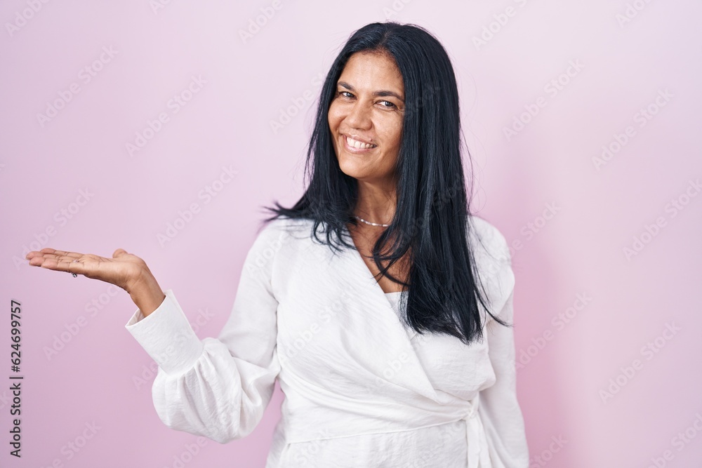 Mature hispanic woman standing over pink background smiling cheerful presenting and pointing with palm of hand looking at the camera. Mature hispanic woman standing over pink background smiling cheerful presenting and pointing with palm of hand looking at the camera.