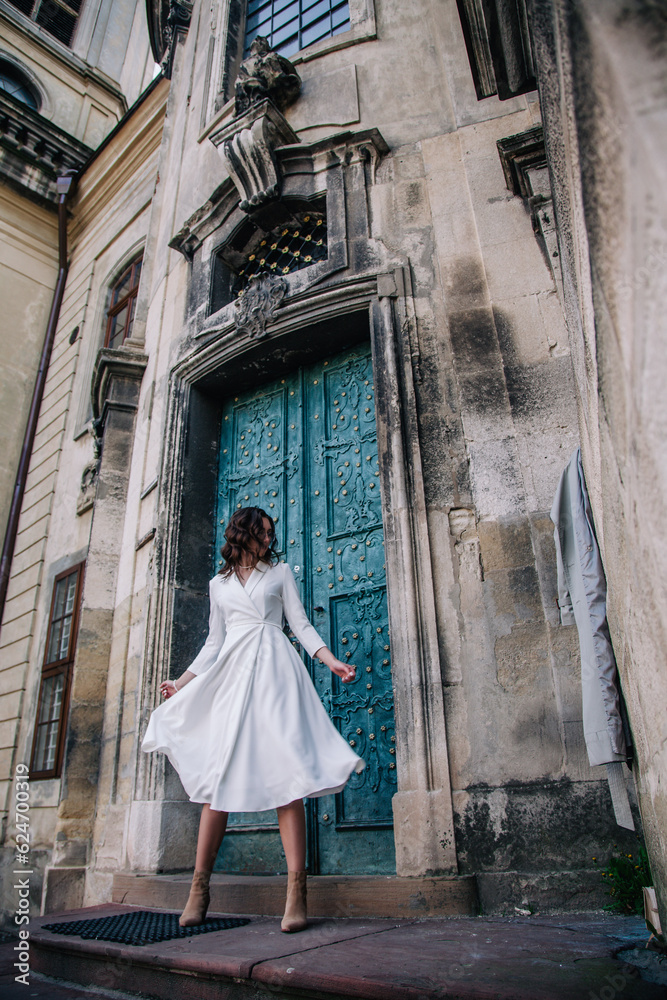 A young, beautiful girl in a white dress and a gray cloak poses near the ancient gray doors of the temple.