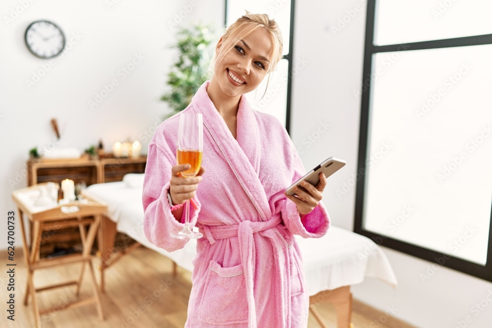 Young caucasian woman wearing bathrobe drinking champagne using smartphone at beauty salon