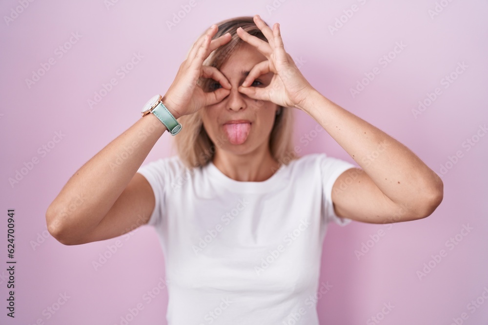 Young blonde woman standing over pink background doing ok gesture like binoculars sticking tongue out, eyes looking through fingers. crazy expression.