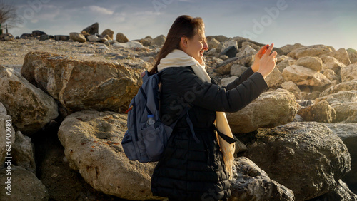 Young woman capturing the beauty of the winter sea by taking pictures with her smartphone while standing on the rocks. Ideal for vacation and travel.
