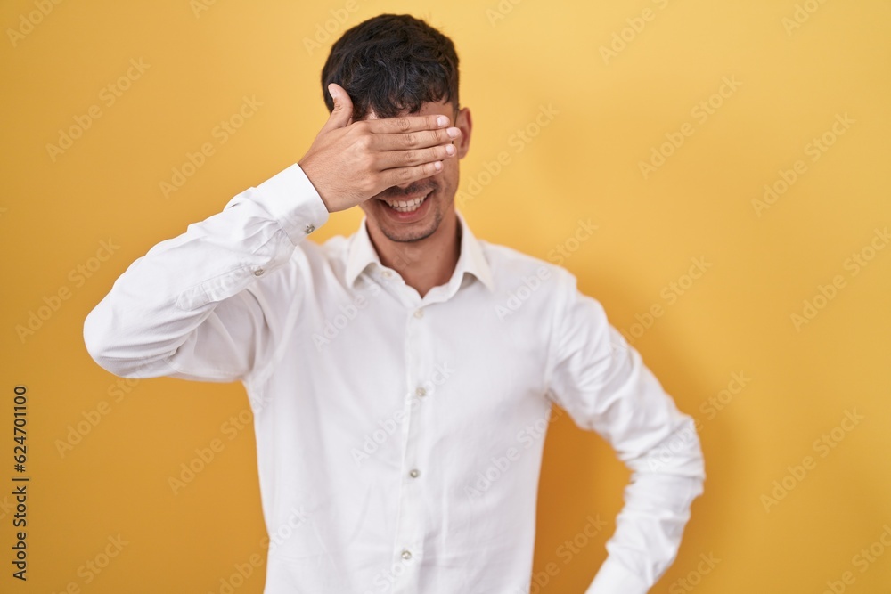 Young hispanic man standing over yellow background smiling and laughing with hand on face covering eyes for surprise. blind concept. Young hispanic man standing over yellow background smiling and laughing with hand on face covering eyes for surprise. blind concept.