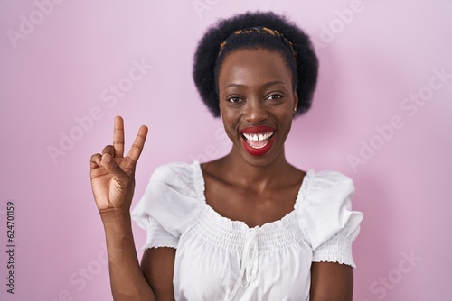African woman with curly hair standing over pink background smiling with happy face winking at the camera doing victory sign with fingers. number two.