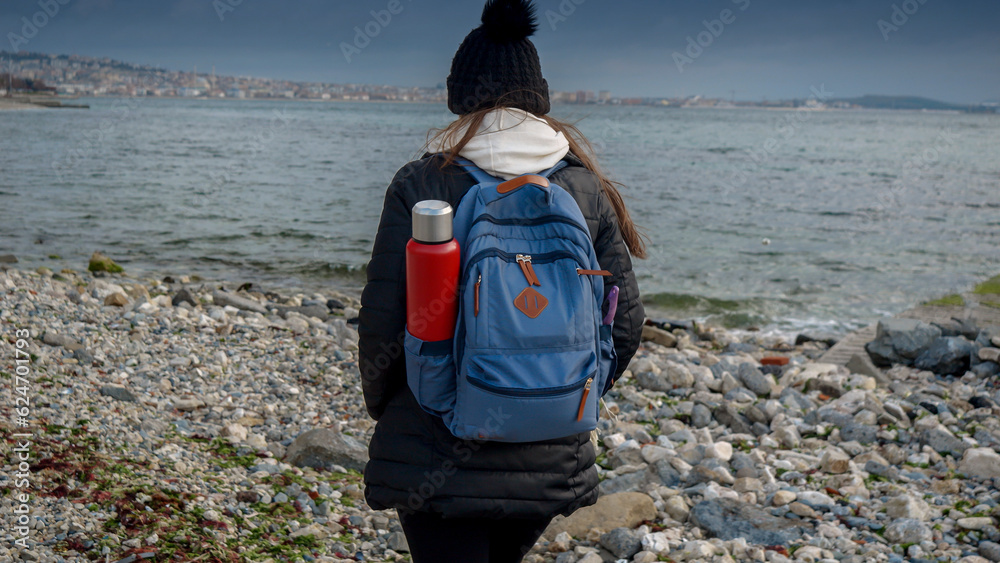 Young female hiker with backpack and thermos walking on the rocky sea beach at cold cloudy and windy day. Concept of hiking adventure, exploration, tourism and journey.