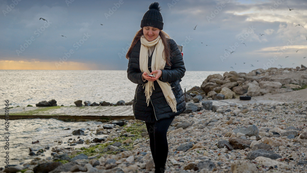 Smiling woman having a walk on the ocean beach at cloudy winter day and picking up sea shells. Concept of hiking, travel, exploration, tourism and journey.
