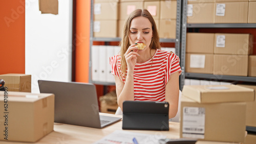 Young blonde woman ecommerce business worker using touchpad eating croissant at office
