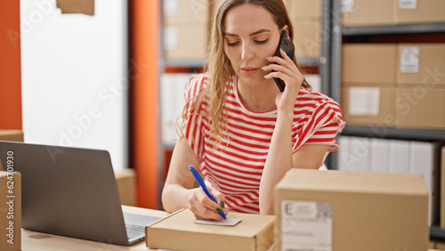 Young blonde woman ecommerce business worker talking on smartphone writing on package at office