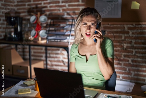 Young beautiful woman working at the office at night speaking on the phone in shock face, looking skeptical and sarcastic, surprised with open mouth