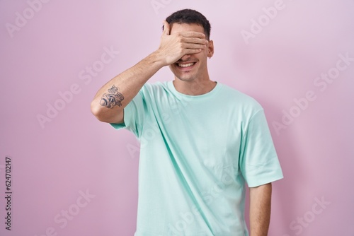 Handsome hispanic man standing over pink background smiling and laughing with hand on face covering eyes for surprise. blind concept.