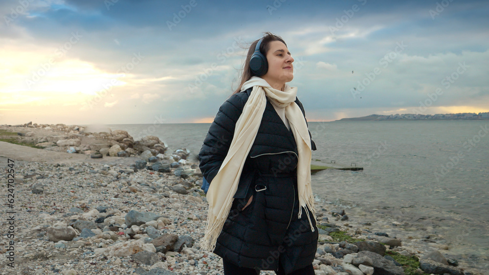 Smiling woman walking along the seashore with her backpack and earphones on. Sense of adventure and wanderlust, making it perfect for use in travel, tourism, and leisure-related content.