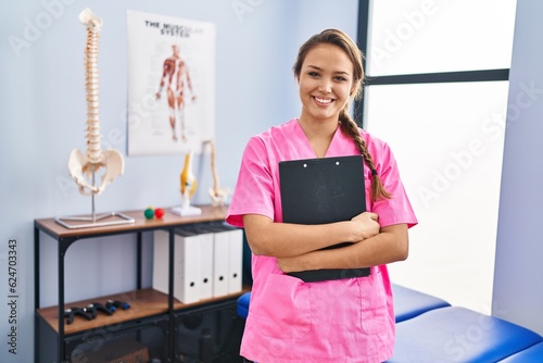 Young beautiful hispanic woman physiotherapist smiling confident holding document at rehab clinic