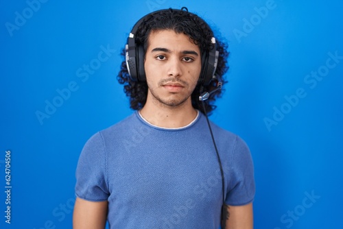 Hispanic man with curly hair listening to music using headphones relaxed with serious expression on face. simple and natural looking at the camera.