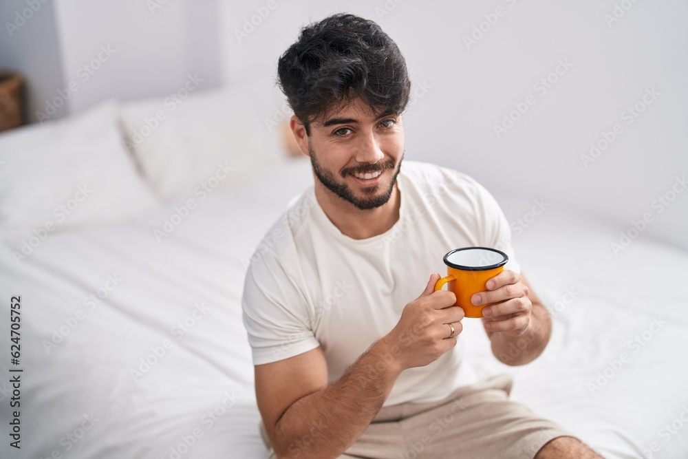Young hispanic man drinking cup of coffee sitting on bed at bedroom Young hispanic man drinking cup of coffee sitting on bed at bedroom