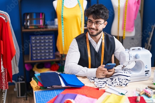 Young hispanic man tailor smiling confident using smartphone at sewing studio