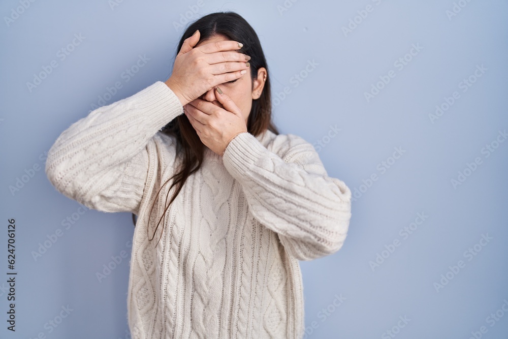 Young brunette woman standing over blue background covering eyes and mouth with hands, surprised and shocked. hiding emotion Young brunette woman standing over blue background covering eyes and mouth with hands, surprised and shocked. hiding emotion