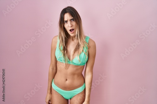 Young hispanic woman wearing bikini over pink background in shock face, looking skeptical and sarcastic, surprised with open mouth
