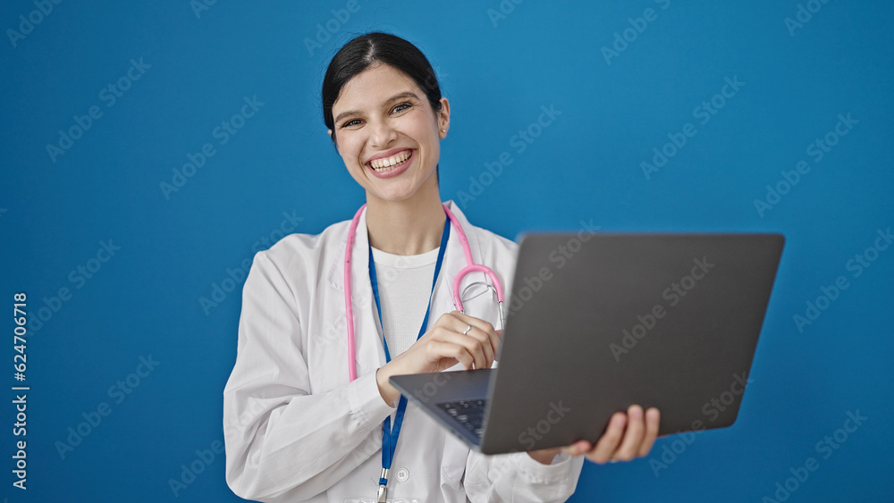 Young beautiful hispanic woman doctor using laptop standing over isolated blue background Young beautiful hispanic woman doctor using laptop standing over isolated blue background